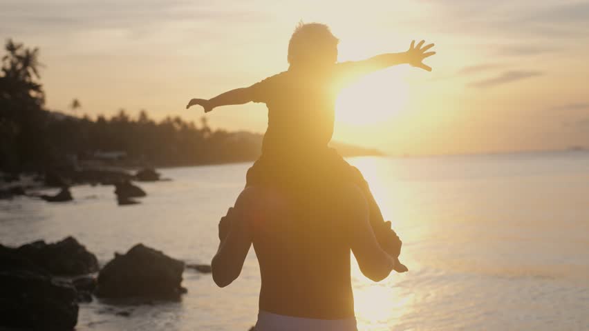 Happy father and son in eyeglasses playing on tropical beach boy rising up hands imitating a flight at wonderful sunset through the shining sun. sow motion. 3840x2160 | Shutterstock HD Video #1008325381