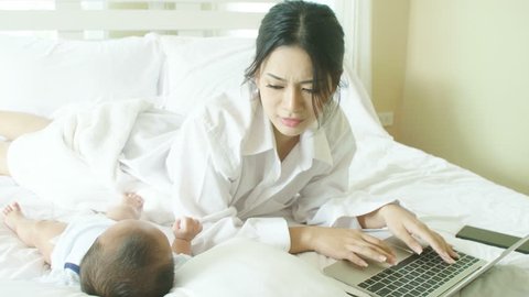Young asian mother with her newborn child working on laptop in bedroom at home.