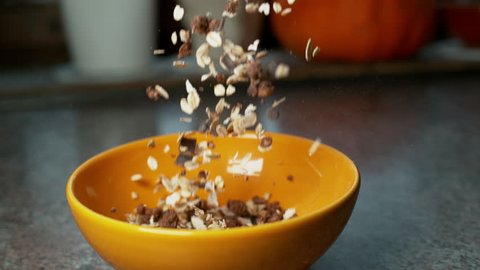 SUPER SLOW MOTION: Nutritious mixture of rolled oats, bran cereal and chocolate pieces land in orange breakfast bowl. Healthy chocolate cereal is being poured into bowl. Nourishing healthy breakfast.