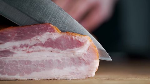 Chef cuts pieces of smoked bacon by sharp knife on the wooden board, cooking meat, meals with meat products, cooking pork