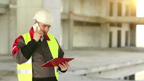 Builder with smartphone at construction site. Foreman with work documentation in yellow hard hat on construction site talks on smartphone. Builder speaks on cell phone on project site