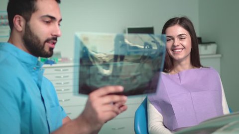 Good-looking doctor demonstrating x-ray to beautiful caucasian patient, young man wearing blue scrubs and holding teeth photograph in right hand, happy woman looking attentively Stockvideó