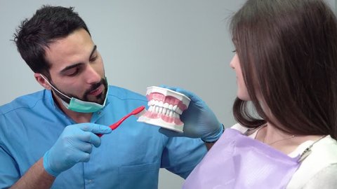 Dentist showing how to clean teeth, handsome doctor demonstrating brushing movements with red toothbrush on dental model, young woman watching him carefully Video Stok