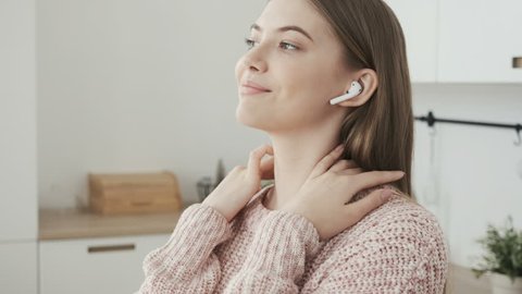 Happy young woman dancing in kitchen and listening to music on wireless earphones. Pretty girl smiling