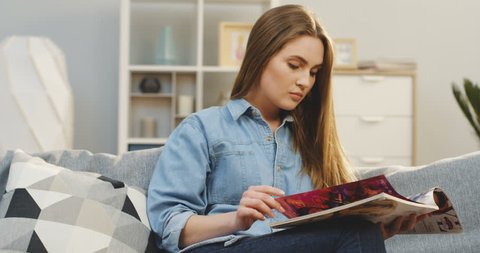 Portrait of the beautiful young woman in jeans shirt reading a magazine while sitting at home on the couch at home. Indoors