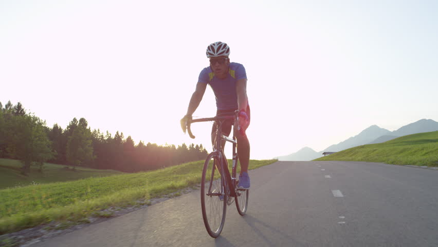 LENS FLARE: Professional biker raising his arms victoriously after winning race. Cheerful biking athlete celebrates victory in quiet countryside. Pro cyclist cheerfully rides road bike with no hands. | Shutterstock HD Video #1008368002