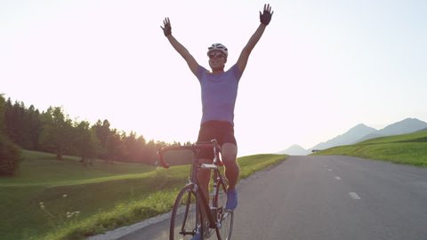 LENS FLARE: Professional biker raising his arms victoriously after winning race. Cheerful biking athlete celebrates victory in quiet countryside. Pro cyclist cheerfully rides road bike with no hands.