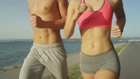 SLOW MOTION, CLOSE UP: Happy Caucasian couple on vacation runs along the sunny ocean road. Cheerful fit girlfriend and boyfriend running together near the beach. 