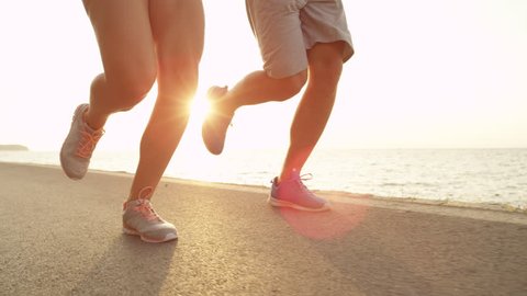 SLOW MOTION, LOW ANGLE, LENS FLARE: Unrecognizable joggers running near the beach in sporty sneakers. Fit couple runs on perfect day at sunny seaside. Unknown people jogging along sunlit walkway.