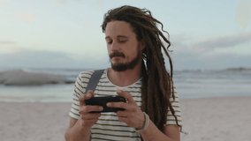 young carefree man with dreadlocks taking photos on beach using phone wearing striped shirt sunset vacation