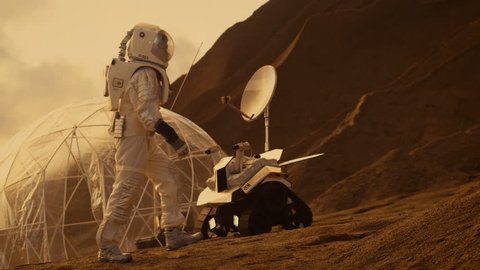 Astronaut Goes Up the Mountain to Explore Red Planet/ Mars. In the Background His Base and AI Powered Rover. Futuristic Colonization Concept. Shot on RED EPIC-W 8K Helium Cinema Camera.