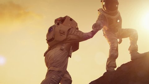 Two Astronauts Climbing Mountain Hill Helping Each Other, Reaching the Top. Helping Hand. Overcoming Difficulties, Important Moment for the Human Race. Shot on RED EPIC-W 8K Helium Cinema Camera.