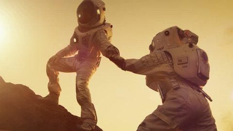 Two Astronauts Climbing Mountain Hill Helping Each Other, Reaching the Top. Overcoming Difficulties, Important Moment for the Human Race. Shot on RED EPIC-W 8K Helium Cinema Camera.