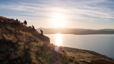 Aerlal view reveal of a group of hikers hiking up steep terrain with lake and golden sunset in the background. 