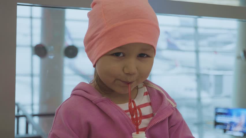 Portrait of a little cute child drinks a juice with drinking straw and looking into the camera at airport with airplanes on the background. Baby waiting flight at departure lounge in slow motion.