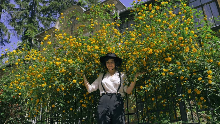 A girl with hat on her head is playfully hiding near the bush with flowers