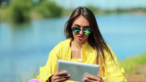 Girl sits near lake and talks via tablet computer. Sportswoman in yellow sports jacket sits near pond and hold in hands tablet pc. Woman in sportswear with tablet pc communicates through videochat