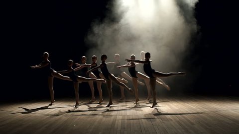 Young teen girls perform ballet on stage in smoke on black background. A choreographed dance of a group of graceful pretty young ballerinas practicing on stage in a classical ballet school.