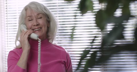 A mature Caucasian woman standing in front of a sunlit window using a landline telephone engaged happily in lively conversation 4k