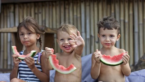 Three preschool children eating watermelon at the beach, smallest child waving hand at someone and smiling