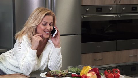 Gorgeous cheerful mature blond haired woman housewife smiling happily talking on the phone at the kitchen preparing to cook dinner culinary cuisine lifestyle leisure healty eating.