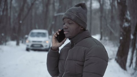 Black man in winter jacket talking on smartphone standing on remote winter road with broken car. Stockvideó