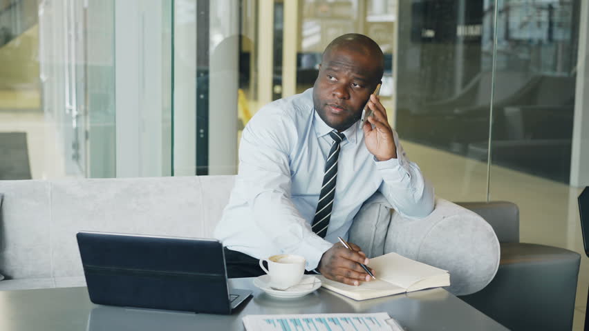 African American businessman talking on smartphone and writing down business information in his notepad sitting in glassy cafe with laptop and coffee on his table.