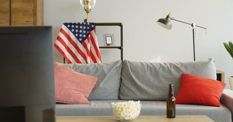 Young Caucasian man coming in the living floor, jumping on the sofa and turning on TV. Cheering up for sport winning with an American flag behind. Popcorn and beer on the table. Indoors