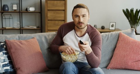 Portrait of the young excited man eating popcorn and changing channels when sitting on the couch at home. Indoor