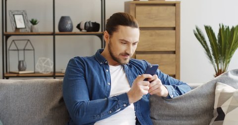 Young attractive man with a tail in the jeans shirt sitting on the sofa in the living room and taping on the smartphone. Inside
