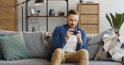 Excited caucasian man in the casual style sitting on the couch in the cozy room and playing games on the smartphone, then angry because loosing. At home. Inside