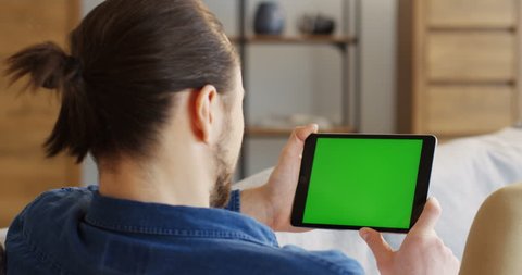 View from the shoulder of young man with a tail holding a tablet computer with a green screen. Chroma key. Close up. Indoor