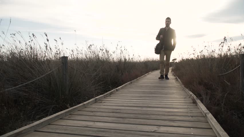 Cheerful adult man walking on pier in dry grass with a laptop bag in sunlight.