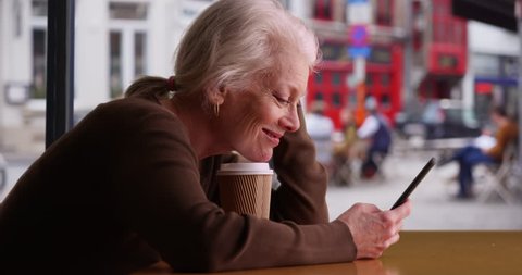 Happy senior woman receives good news via text while waiting in coffee shop. Smiling retiree using smartphone in cafe and drinking coffee. 4k