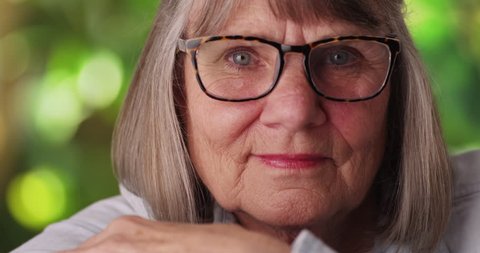 Close-up of somber elderly woman wearing eyeglasses outside. Close view of relaxed senior lady looking at camera in lush outdoor setting. 4k