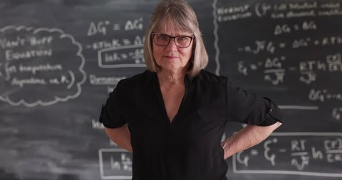 Old senior woman teacher with serious attitude posing in front of chalkboard. Charming mature woman with arms on her waist looking at camera . 4k