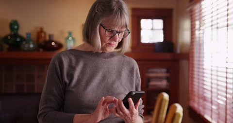Serious senior woman using phone reading tragic news standing in living room. Somber lady reading e-mail on cell phone looking concerned in indoor setting. 4k