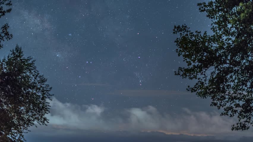 Milky Way time lapse at night in the northern hemisphere with rolling clouds moving in the opposite direction as the stars as trees blow in the wind on each side.