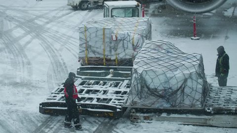 Two airport laborers attempt to re a large pallet of goods from a conveyor belt and put them onto a loading truck in a snow storm
