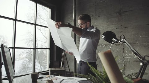Bearded man (hipster) standing faces to panoramic window and looks at blueprints. Young architect in stylish loft office works with documents. Creative architect thinking on the big drawings.