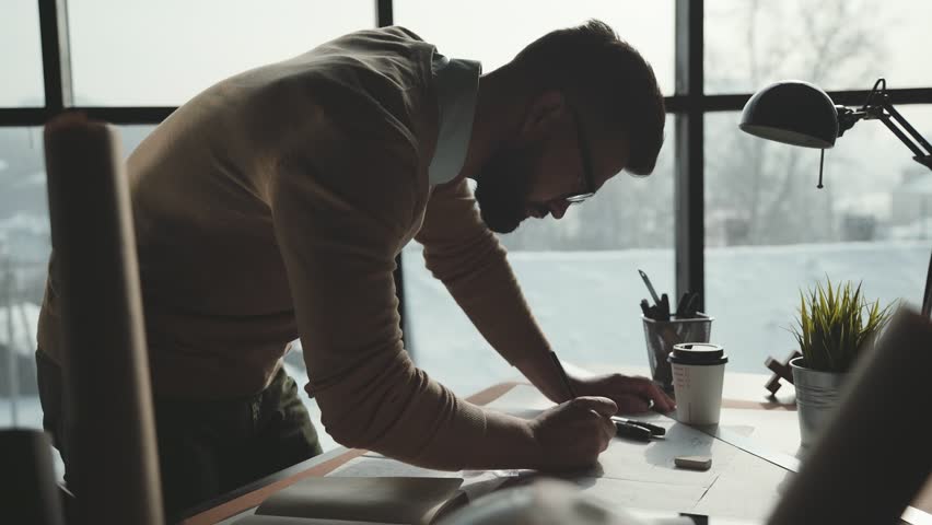 Bearded man in glasses makes sketches in front of large panoramic window. Engineer works in bright office with a large window, concentrates and draws blueprints. Workplace of an architect or designer | Shutterstock HD Video #1008551881