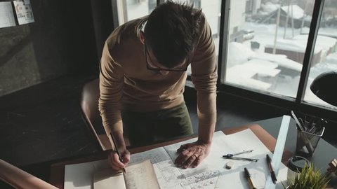 Hipster engineer works with blueprint. Shoot from above. Young architectural engineer working in office. Bearded man in glasses makes sketches. Loft style, minimalistic interior, drawings on the table