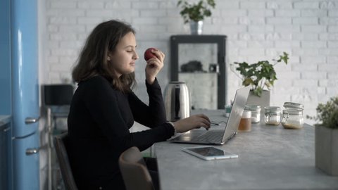 Teenage girl using laptop and eating apple sitting by table at home 
