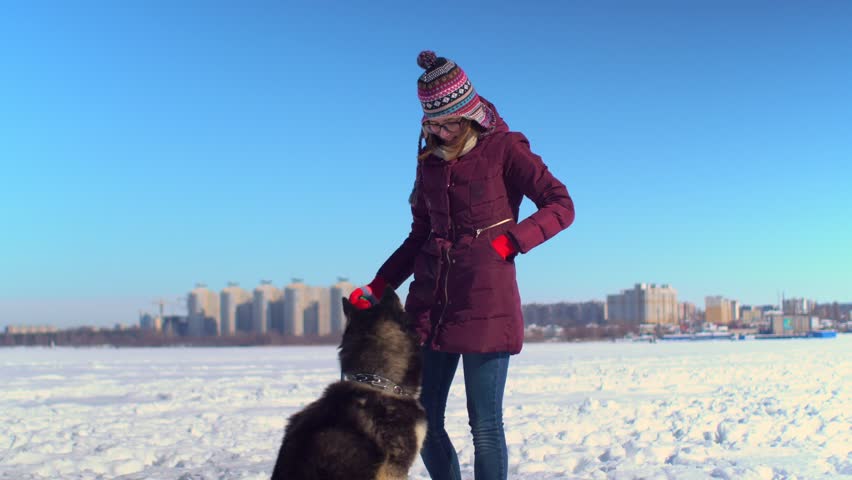 Attractive girl playing with her Husky dog on frozen river against background of cityscape.