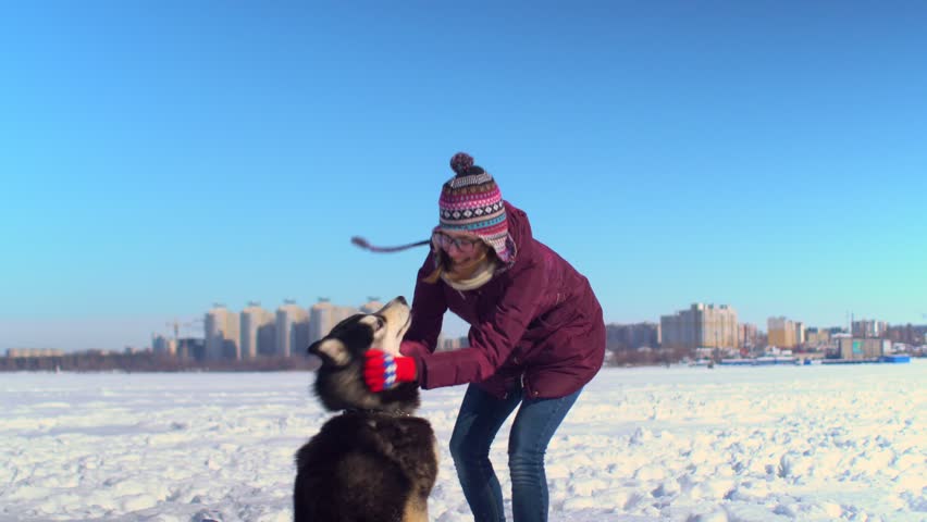 Attractive young woman playing with her Husky dog on frozen river against background of cityscape.