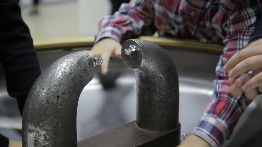 Children are experimenting with device at scientific exhibition in room. Curious boys are testing new invention on table at science fair. Curious kids attach nuts to metal object with its own magnetic