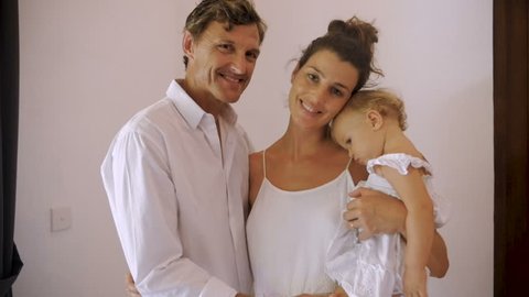 Portrait of a beautiful family including a mother, a sleepy tired baby girl, and the father standing, looking at the camera in slow motion