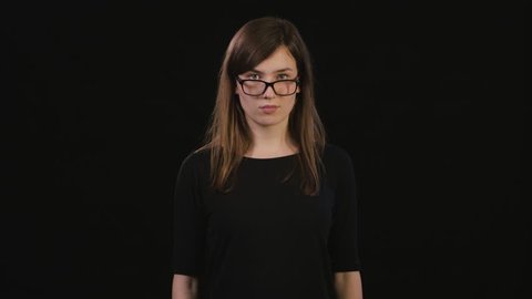 A beautiful young lady touches her black glasses with her finger against a black background. Medium Shot