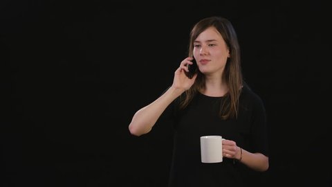 A beautiful young lady talking on the phone and holding a white cup against a black background. Medium Shot