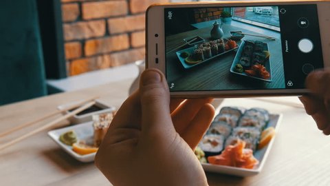 Hands of a teenager make a photo of food on a smartphone. A set of sushi rolls from Japanese cuisine on the background of a porcelain teapot for soy sauce and saucer in a stylish cafe